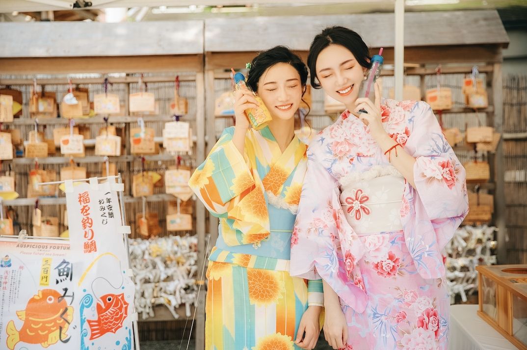 明るい黄色と淡いピンクの浴衣を着た女性二人が、色柄の違いを楽しみながら神社で撮影する様子