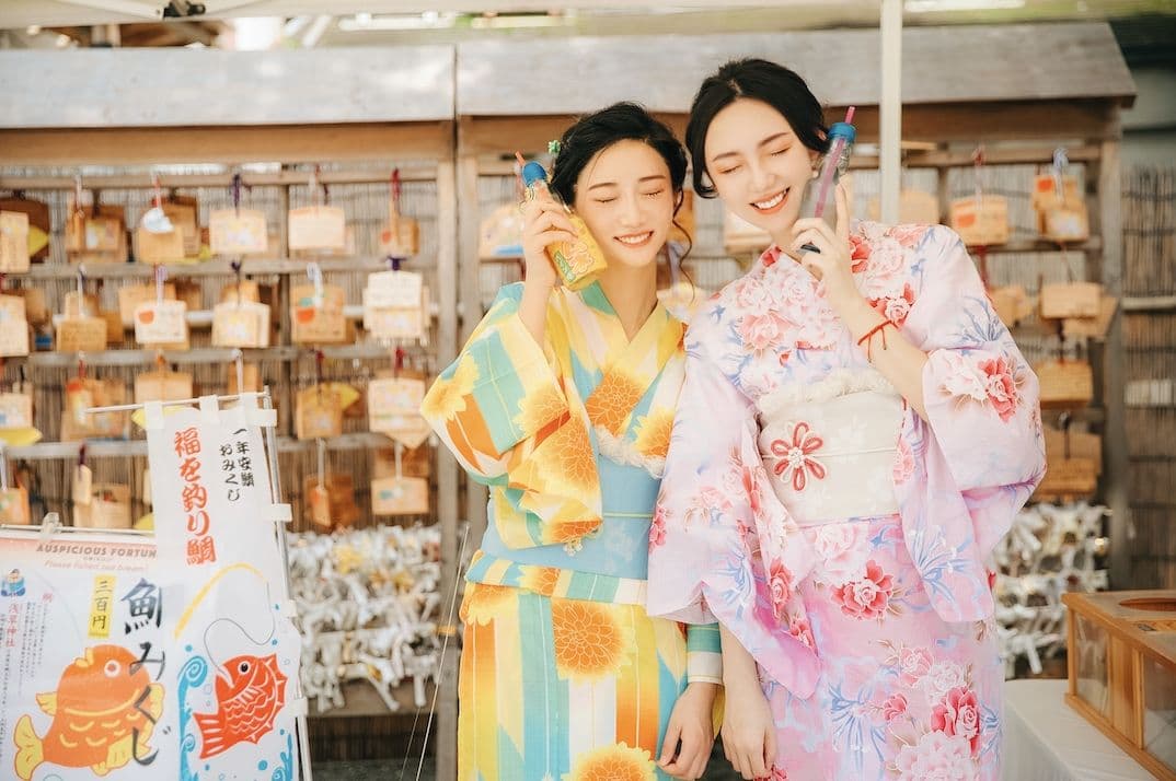 明るい黄色と淡いピンクの浴衣を着た女性二人が、色柄の違いを楽しみながら神社で撮影する様子